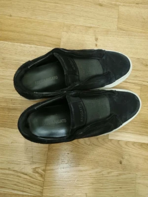 Svarta slip-on sneakers från Axel Arigato - Snygga svarta slip-on sneakers från Axel Arigato med vit sula och elastiskt band över vristen. Ovandelen är i mocka och textil, vilket ger en stilren och modern look. Perfekta för dig som gillar minimalistisk design och enkelhet.