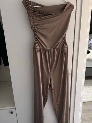 Beige off shoulder jumpsuit - Snygg beige jumpsuit med off shoulder-design och draperad överdel. Tillverkad i ett glansigt, stretchigt material som ger en smickrande passform. Perfekt för dig som vill sticka ut med en stilren look.