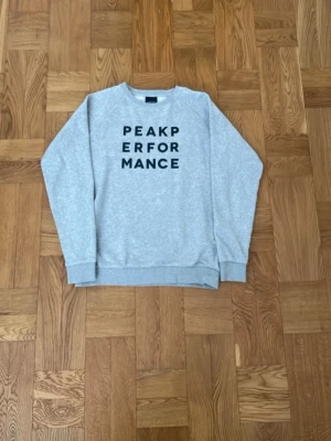 Grå sweatshirt från Peak Performance - Snygg grå sweatshirt från Peak Performance med svart tryck på bröstet. Tröjan har rund halsringning, ribbade muddar. Oanvänd i väldigt bra skick