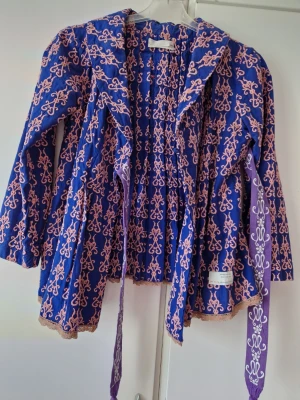 Blå omlottjacka med broderi - Unik blå omlottjacka från Odd Molly med rosa broderat mönster och dekorativa band. Jackan har lång ärm, volangkant och knytband i midjan. Perfekt för dig som vill sticka ut med färg och mönster.