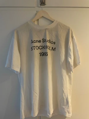Vit Acne Studios t-shirt Stockholm - Vit t-shirt från Acne Studios med svart text 'Acne Studios STOCKHOLM 1996' på bröstet. Klassisk passform med rund hals och korta ärmar. Tillverkad i mjuk bomull, perfekt för en clean och enkel stil.