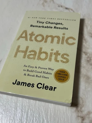 Atomic Habits av James Clear - Säljer boken 'Atomic Habits' av James Clear. Pocketbok med ljusgul framsida och guldiga bokstäver. Perfekt för dig som vill lära dig mer om vanor och personlig utveckling. Texten är på engelska och boken är tryckt på vitt papper.