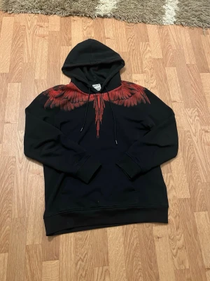 Marcelo burlon  - Säljer en svart hoodie från Marcelo Burlon med ikoniskt rött vingtryck över axlar och bröst. Hoodien har huva med dragsko och ribbade muddar. Perfekt för dig som vill sticka ut med en unik och trendig design.