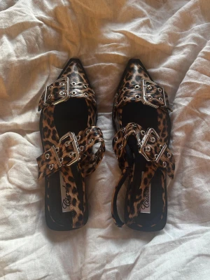 Leopardmönstrade ballerinaskor med spännen - Snygga ballerinaskor från Chiara Ferragni i leopardmönstrat material. Skorna har spetsig tå, dubbla breda remmar med silvriga spännen och öljett-detaljer. Sulan är svart och platt. Perfekta för dig som vill sticka ut med din stil.