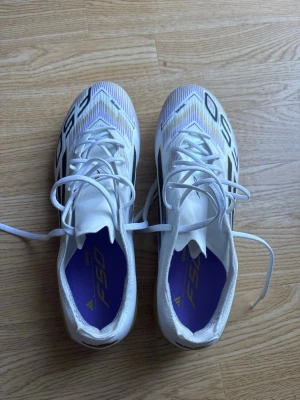 Adidas F50 vita fotbollsskor - Säljer ett par Adidas F50 fotbollsskor i vitt med svarta och guldiga detaljer. Skorna har klassisk snörning. Gräsdobbar i storlek 41. Endast använda en gång, så är i utmärkt skick!