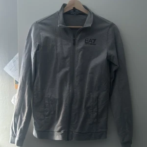 Grå tröja från Emporio Armani - Säljer en stilren grå tröja från Emporio Armani med dragkedja framtill. Tröjan har en enkel design med EA7-logga på bröstet och långa ärmar. Perfekt för en casual look.