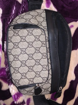 Beige axelväska från Gucci - Snygg beige axelväska från Gucci med klassiskt GG-mönster och svarta detaljer i läder. Väskan har en framficka med dragkedja och justerbar axelrem. Perfekt för att bära det viktigaste med stil.
