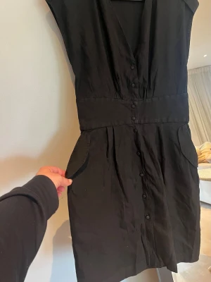 Sandro klänning  - Stilren svart linne klänning med knappar framtill och markerad midja. Klänningen har korta ärmar, v-ringning och två fickor framtill. Passar perfekt för dig som gillar en klassisk look. Sval klänning för sommaren