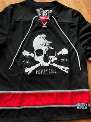 Limiterad Mötley Crüe Jersey Tour 2015 - Svart t-shirt med vit dödskalle och korslagda benknotor framtill, samt texten 'Mötley Crüe 1981-2015'. Röd och grå rand nertill och snörning vid halsen. Perfekt för dig som gillar rock och bandmerch. 
