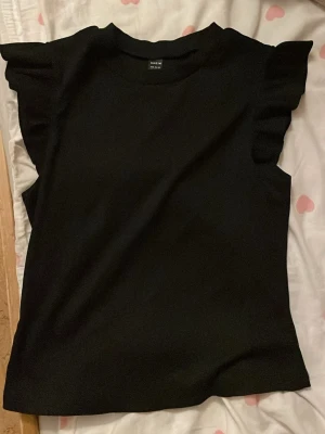 Svart volangärmad topp från Shein - Snygg svart topp från Shein med volangdetaljer på axlarna. Ärmlös modell med rund halsringning och rak passform. Perfekt att matcha med jeans eller kjol för en trendig look.