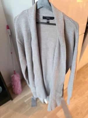 Ljusgrå kofta från Pieces - Mysig ljusgrå stickad kofta från Pieces med öppen front och knytskärp i midjan. Koftan har lång ärm och bred krage, perfekt för lager-på-lager-stil. Enkel och stilren design som passar till det mesta.