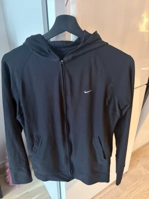 Svart Nike hoodie med dragkedja - Svart hoodie från Nike med dragkedja framtill och två fickor. Klassisk design med liten vit Nike-logga på bröstet och huva. Perfekt för en sportig och avslappnad stil. Tillverkad i mjukt material som känns skönt mot huden.