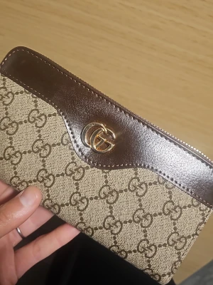 Beige och brun plånbok - Snygg plånbok  i beige canvas med klassiskt GG-mönster och bruna läderdetaljer. Guldigt GG-emblem på framsidan och dragkedja runt om. Perfekt för dig som vill ha en lyxig och stilren accessoar.