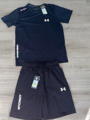 Svart träningsset från Under Armour - Snyggt svart träningsset från Under Armour med t-shirt och shorts. Båda plaggen har logga och tryck i vitt. T-shirten har rund hals och kort ärm, shortsen har resår och dragsko i midjan. Perfekt för gym eller löpning.