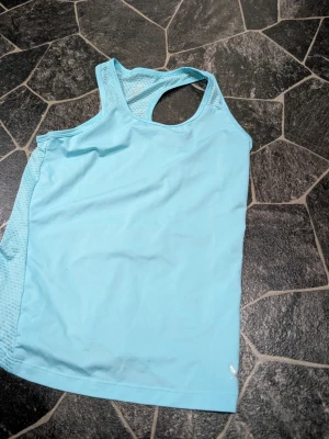 Ljusblått sportlinne i mesh stl 146-152 - Snyggt ljusblått sportlinne med brottarrygg och meshpaneler på baksidan för extra ventilation. Perfekt för träning och aktiva dagar. Tillverkat i lätt och snabbtorkande material som håller dig sval under passet.