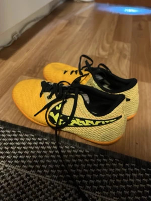 Gula Nike fotbollsskor inomhus - Säljer ett par gula Nike fotbollsskor för inomhusbruk med svart sula och snörning. Skorna har ett coolt mönster på sidan och är tillverkade i ett ventilerande meshmaterial. Perfekta för snabba vändningar på planen och grymt grepp på inomhusgolv.