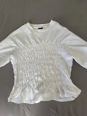 Vit smockad topp från Gina Tricot - Vit topp från Gina Tricot med smockad front och lätt volang nertill. Modellen har rund halsringning och korta ärmar. Tillverkad i mjuk bomull🤍