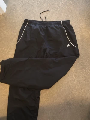 Svarta mjukisbyxor från Adidas  - Säljer ett par svarta mjukisbyxor från Adidas med vita detaljer vid fickorna och klassisk logga på ena benet. Byxorna har dragsko i midjan och är tillverkade i ett lätt och ventilerande material med meshfoder. Perfekta för chill eller träning.