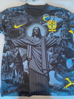 Brasil Nike fotbollströja svart/grå - Unik svart och grå Brasilien fotbollströja från Nike med stort tryck av Kristusstatyn och detaljerade illustrationer. Gult Nike-märke och Brasil-logga på bröstet. Rund halsringning och korta ärmar. Perfekt för dig som vill sticka ut på planen.
