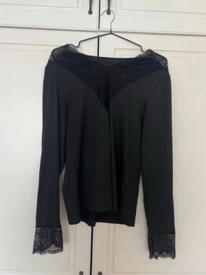 Svart blus med spetsdetaljer - Stilren svart blus med långa ärmar och vackra spetsdetaljer vid halsringning och ärmslut. Blusen har en elegant, något lös passform och ger en sofistikerad look. Perfekt för dig som gillar en klassisk men trendig stil.