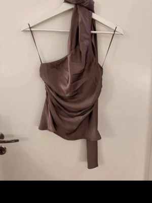 Brun/lila satin one shoulder topp Zara - Snygg brun one shoulder topp från Zara i glansigt satinmaterial. Toppen har en lång knytband-detalj vid axeln och draperad effekt framtill. Perfekt för dig som vill sticka ut med en trendig och elegant look.