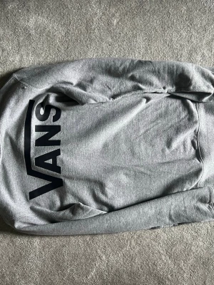 Grå sweatshirt från Vans - Snygg grå sweatshirt från Vans med stort svart logotryck på ryggen. Tröjan har rund halsringning och långa ärmar. Perfekt för en avslappnad streetstyle-look.