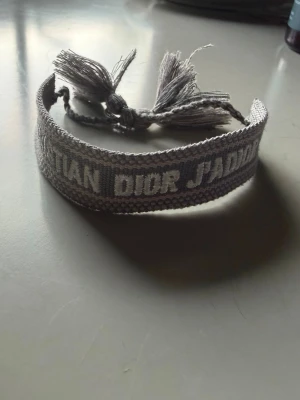 Grått broderat armband från Dior - Snyggt grått armband från Dior med broderad text 'Christian Dior J'Adior'. Armbandet är gjort i tyg och har tofsar i ändarna för en cool och trendig look. Perfekt accessoar för att lyfta din outfit.
