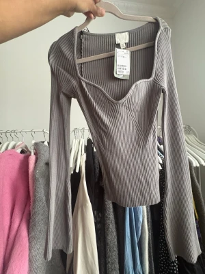 Grå ribbad långärmad topp H&M - Trendig grå ribbad topp från H&M med lång ärm och snygg v-ringad urringning. Figurnära passform och markerade sömmar framtill ger en modern look. Perfekt att styla med jeans eller kjol för en cool outfit.