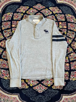 Grå långärmad topp Abercrombie - Grå långärmad topp från Abercrombie & Fitch med knappar vid halsen och mörkblå broderad älg på bröstet. Två mörkblå ränder på ena ärmen ger en sportig touch. Mjuk och skön bomullskvalitet.