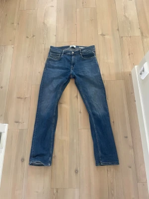 Blå raka jeans från Replay - Klassiska blå jeans från Replay med rak passform och fem fickor. Jeansen har en snygg tvätt och subtila slitningar för en avslappnad look. Tillverkade i denim med knapp- och dragkedjegylf. Perfekta för en stilren vardagsstil.