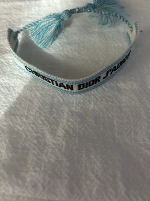 Christian Dior J'adior blått tygarmband - Snyggt armband från Christian Dior i ljusblått och vitt tyg med texten 'J'adior'. Justerbar knytning med tofsar i ändarna. Perfekt accessoar för att lyfta din outfit med en lyxig touch och trendig vibe.