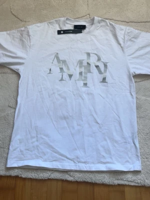 Vit t-shirt från Amiri med logga - Vit t-shirt från Amiri med stor, stilren logga framtill i silver. Klassisk passform och rund halsringning. Perfekt för dig som gillar minimalistisk design och vill ha ett trendigt plagg i garderoben.