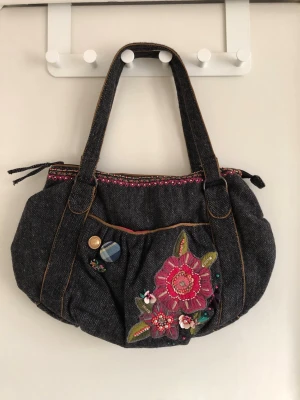 Handväska med broderi - Unik handväska i mörkgrå med två handtag, dekorerad med färgglatt broderi och applikationer i form av blommor och knappar på framsidan. Väskan har kontrastsömmar och dragkedja upptill. Perfekt för dig som gillar en kreativ och personlig stil.