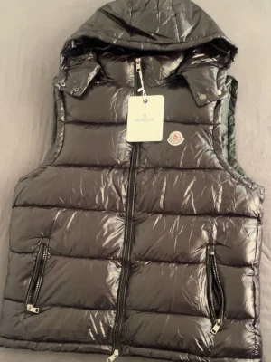 Svart dunväst från Moncler med huva - Snygg svart dunväst från Moncler med glansig finish, avtagbar huva och två dragkedjefickor framtill. Västen har Moncler-logga på bröstet och stängs med dragkedja. Perfekt för lager-på-lager och streetstyle.