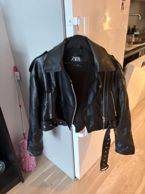 Svart skinnjacka från Zara - Snygg svart skinnjacka från Zara med klassisk bikerstil, silvriga dragkedjor, bred krage och bälte i midjan. Jackan har axelklaffar och detaljerade sömmar som ger en cool look. Perfekt för dig som vill ha en edgy stil.