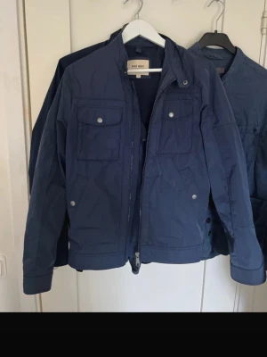 Mörkblå vindjacka från East West - Snygg mörkblå vindjacka från East West med dragkedja framtill och flera fickor med tryckknappar. Jackan har en klassisk passform och är tillverkad i ett lätt och slitstarkt material, perfekt för blåsiga dagar. Skick 9/10