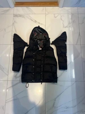 Svart dunväst med avtagbara ärmar - Snygg svart dunväst från Burberry med avtagbara ärmar och stor huva fodrad i klassiskt rutigt tyg. Västen har knappar och dragkedja framtill samt dragsko nertill för justerbar passform. Perfekt för lager-på-lager och streetstyle.