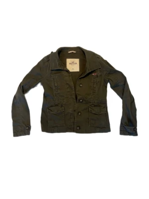 Grön utilityjacka från Hollister - Snygg grön utilityjacka från Hollister med klassisk krage, stora fickor framtill och knappstängning. Jackan har långa ärmar och detaljerade sömmar samt ett diskret Hollister-märke på bröstet. Perfekt för lager-på-lager och streetstyle. Storlek M men sitter som S