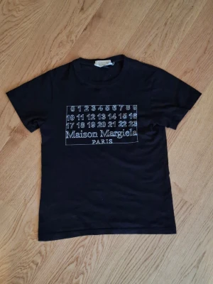 Maison Margiela svart t-shirt - Svart t-shirt från Maison Margiela med deras ikoniska siffror och logga tryckt i vitt på bröstet. Klassisk passform med rund hals och korta ärmar. Tillverkad i mjuk bomull som känns skön mot huden. Storlek 160 - 170