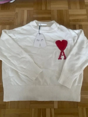 Vit stickad tröja från Ami Paris - Vit stickad tröja från Ami Paris med röd hjärtlogga och bokstaven A på bröstet. Tröjan har ribbstickade muddar vid ärmslut, hals och nederkant. Klassisk passform och rund halsringning. Perfekt för en stilren och trendig look.