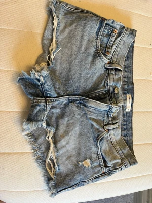 Blå jeansshorts  - Säljer ett par blå jeansshorts från Gina tricot med råa fransade kanter och slitningar. Klassisk femficksmodell med knapp och dragkedja framtill. Perfekta för sommarens alla äventyr!