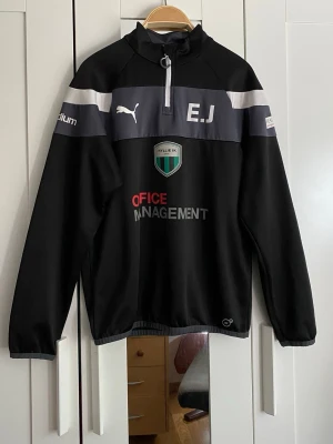Svart träningströja  - Svart långärmad träningströja från Puma med halv dragkedja, klubbmärke för Hyllie IK, tryckta initialer 'E.J' och sponsorer. Tröjan har grå och vita detaljer samt elastiska muddar. Perfekt för fotbollsträning eller uppvärmning.