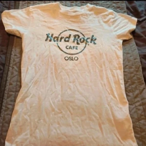 Vit Hard Rock Cafe Oslo t-shirt - Vit Hard Rock Cafe Oslo t-shirt  Vit t-shirt från Hard Rock Cafe Oslo med glittrigt svart tryck på bröstet. Klassisk passform med rund hals och korta ärmar. Tillverkad i 100% bomull, perfekt för dig som gillar ikoniska bandtröjor och vill sticka ut lite extra.  Kan även hämtas I Partille  E1