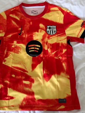 Barcelona matchtröja röd/gul Nike - Barcelona matchtröja i röd och gul tie-dye design med klubbmärke, Nike-logga och Spotify-sponsor på bröstet. Tillverkad i lätt och ventilerande material, perfekt för fotboll eller träning. UNHCR-logga på ryggen. Snygg och unik stil för fans.