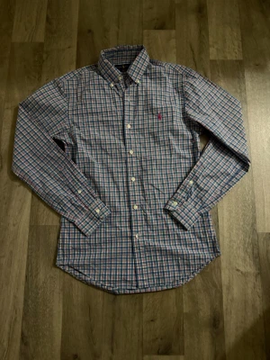 Rutig skjorta från Ralph Lauren - Snygg rutig skjorta från Ralph Lauren i blått, rosa och vitt. Klassisk button-down krage, långa ärmar och broderad logga på bröstet. Perfekt för dig som gillar preppy stil och vill sticka ut lite extra.