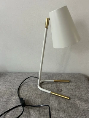 Vit bordslampa med mässingsdetaljer - Modern bordslampa i vit metall med stilrena mässingsdetaljer på fot och arm. Lampan har en konformad skärm och svart sladd med strömbrytare. Perfekt som skrivbordslampa eller läslampa.