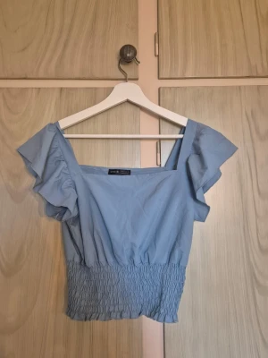 Ljusblå volangtopp - Söt ljusblå topp med breda axelband och volangdetaljer vid ärmarna. Toppen har smockad midja som ger en snygg passform och croppad längd. Perfekt för varma dagar och enkel att styla till jeans eller kjol.    Alla kläder rollas innan dom skickas hem då det finns katt i hemmet🐈 Jag stryker och tvättar INTE innan jag skickar det får du stå för själv så det blir som du vill ha det🤗