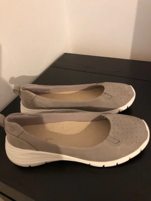 Grå ballerinaskor från Hush Puppies - Snygga grå ballerinaskor från Hush Puppies med vit sula och perforerat mönster på tån. Skorna har en sportig design med elastiska detaljer och mjuk innersula för extra komfort. Perfekta för dig som gillar en stilren och bekväm look.