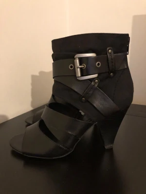 Svarta pumps med spänne och remmar - Snygga svarta pumps med öppen tå, bred klack och flera dekorativa remmar i äkta läder. Skorna har ett stort metallspänne och detaljer i både tyg och läder. Perfekta för dig som gillar en edgy och modern stil.