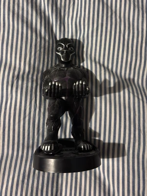 Black Panther figurhållare Marvel - Cool Black Panther figurhållare från Marvel i svart plast med lila detaljer. Figuren står på en rund bas och har utsträckta armar för att hålla till exempel en handkontroll eller mobil. Perfekt för Marvel-fans som vill ha något unikt till skrivbordet.
